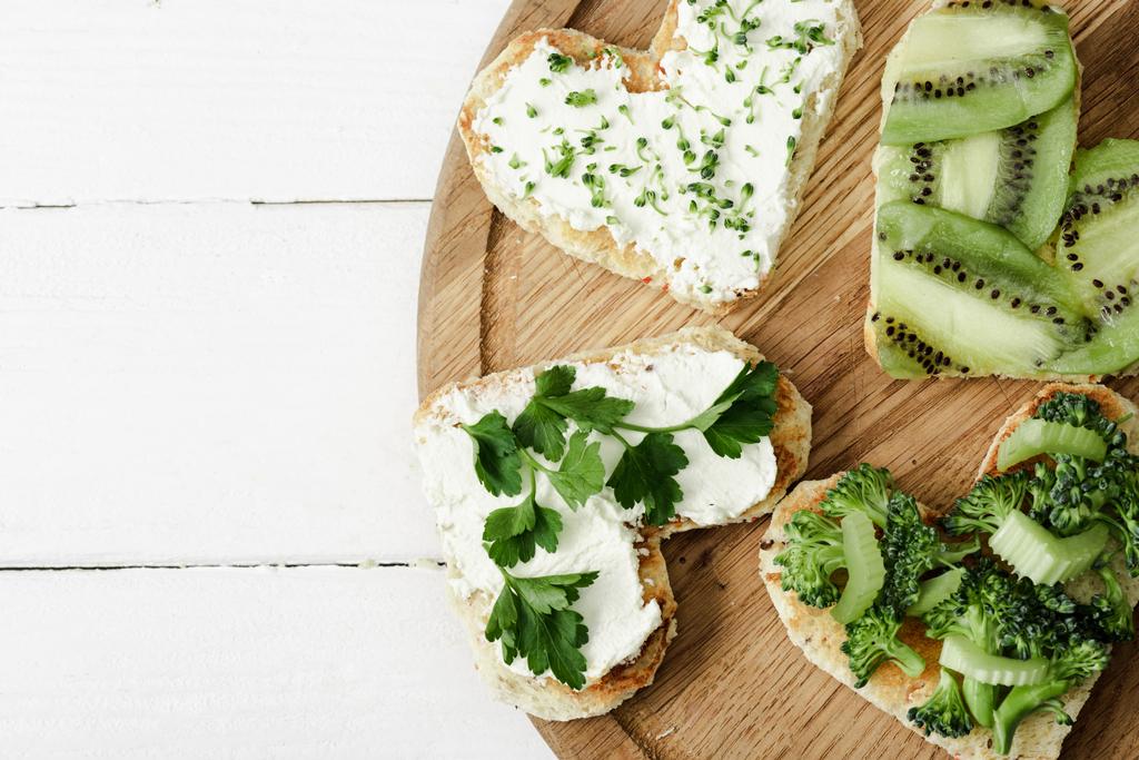 vrchní pohled na srdíčko ve tvaru canape s krémovým sýrem, brokolice, microgreen, petržel a kiwi na palubě na bílém dřevěném povrchu - Fotografie, Obrázek