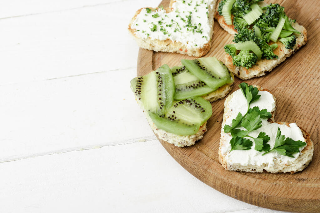 heart shaped canape with creamy cheese, broccoli, microgreen, parsley and kiwi on board on white wooden surface - Photo, Image