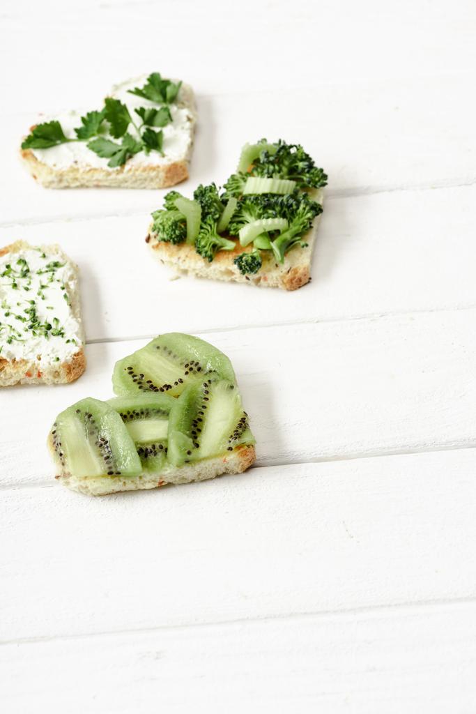 tartina a forma di cuore con formaggio cremoso, broccoli, microverde, prezzemolo e kiwi su superficie di legno bianca
 - Foto, immagini