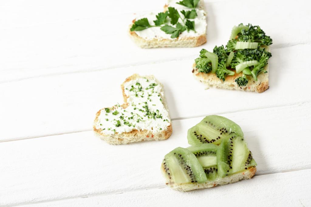 herzförmiger Canape mit cremigem Käse, Brokkoli, Mikrogrün, Petersilie und Kiwi auf weißer Holzoberfläche - Foto, Bild
