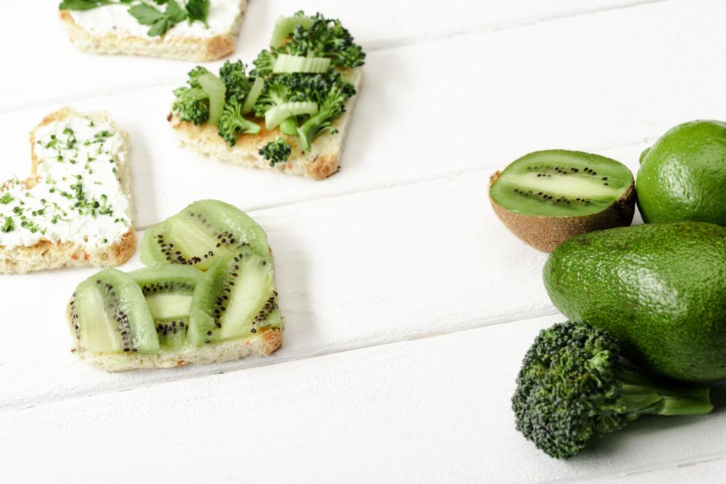střapec ve tvaru srdce s krémovým sýrem, brokolicí, mikrozelení, petrželkou a kiwi v blízkosti zeleného ovoce a zeleniny na bílém dřevěném povrchu - Fotografie, Obrázek