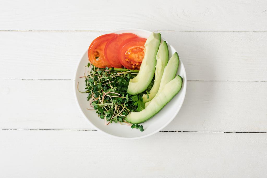 Draufsicht auf Tomate, Avocado und Microgreen in Schale auf weißer Holzoberfläche - Foto, Bild