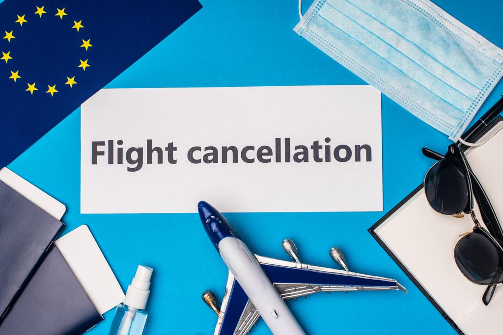 Top view of card with flight cancellation lettering near flag of european unity, toy airplane and medical mask on blue background - Photo, Image