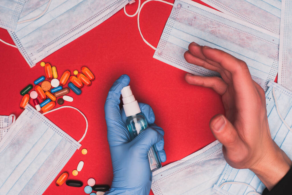 Kırmızı yüzeyde ilaç ve tıbbi maskelerin yanında bir şişe el dezenfektanı tutan adam görüntüsü. - Fotoğraf, Görsel