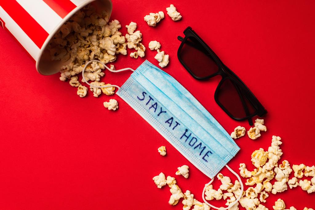 Bovenaanzicht van medisch masker met verblijf thuis belettering in de buurt van zonnebril en popcorn op rode achtergrond - Foto, afbeelding