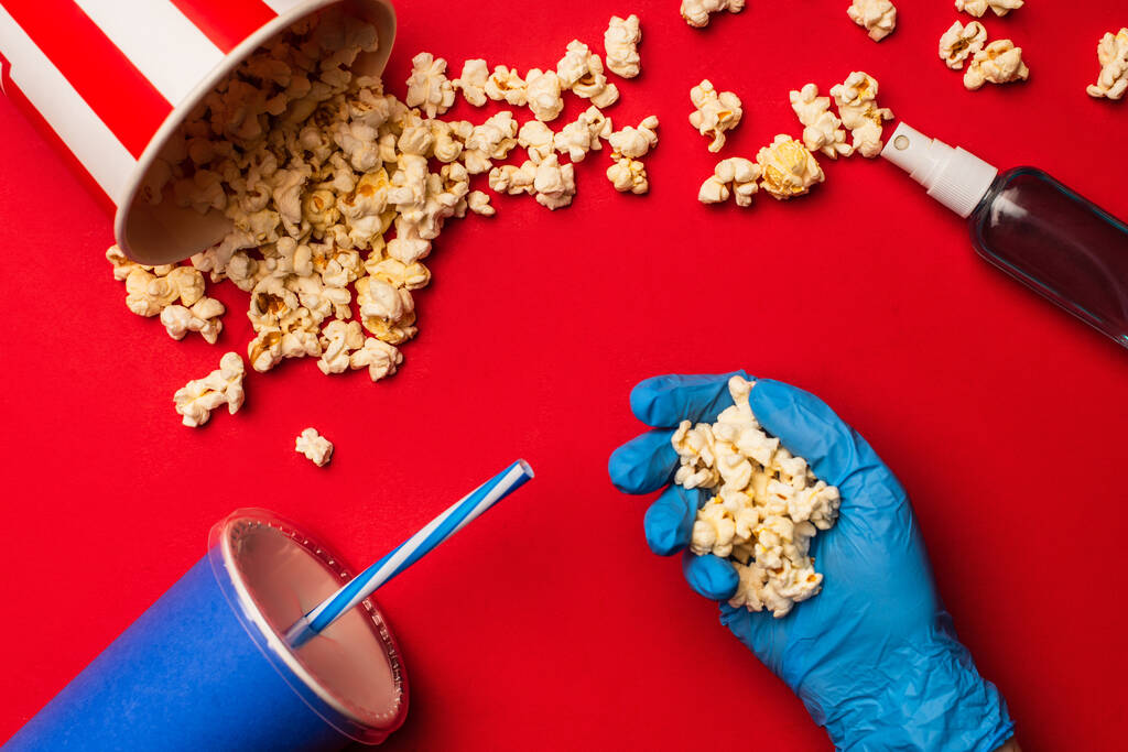 Top view of person in latex glove holding popcorn near hand sanitizer and paper cup on red background - Photo, Image