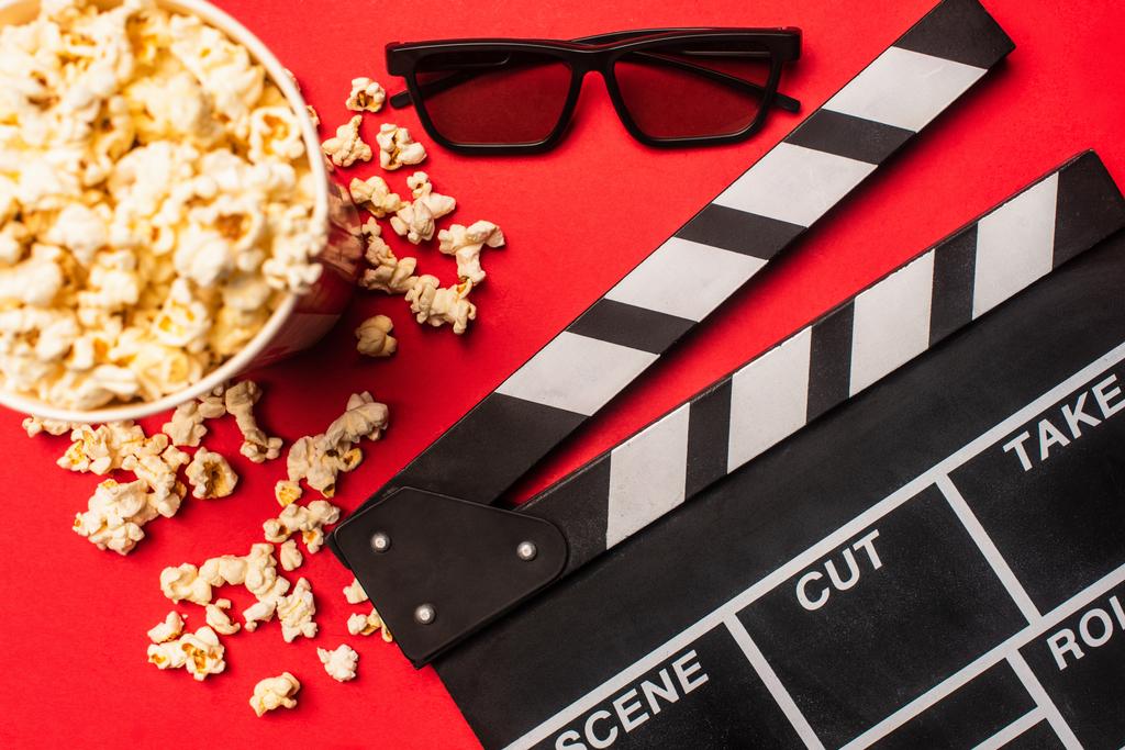 Bovenaanzicht van clapperboard bij emmer met popcorn en zonnebril op rode achtergrond - Foto, afbeelding