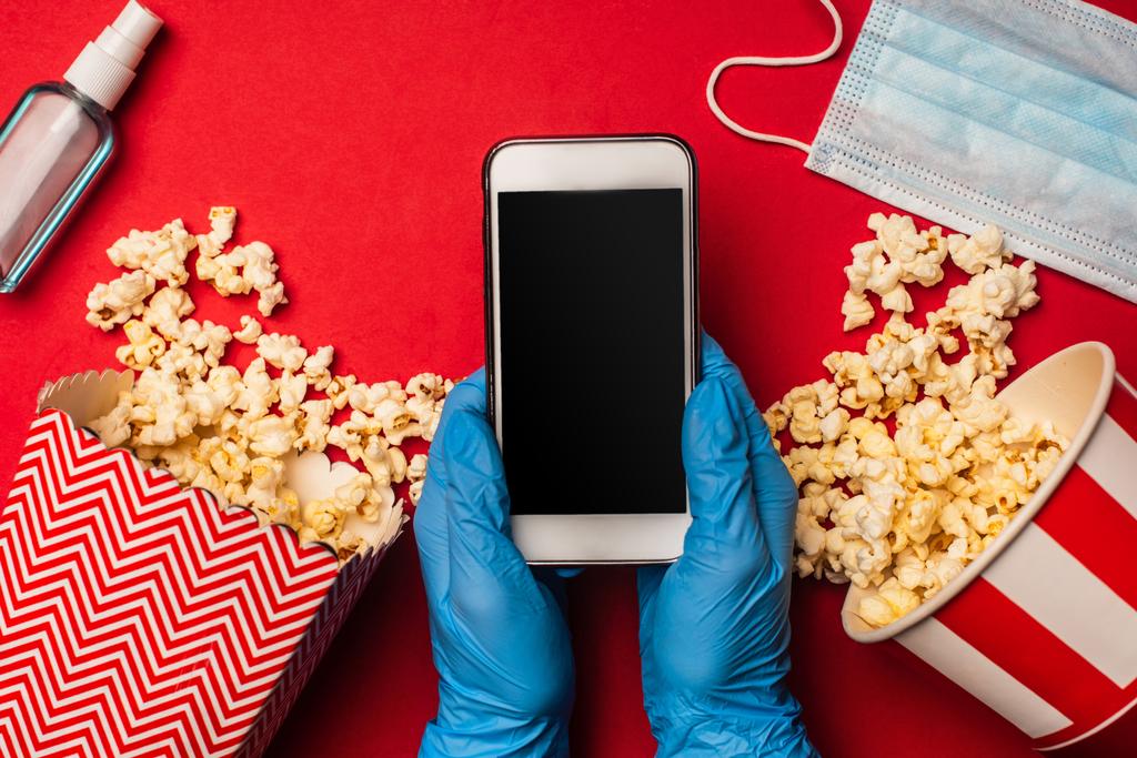 Horní pohled na osobu držící smartphone s prázdnou obrazovkou v blízkosti lékařské masky a popcorn na červeném povrchu - Fotografie, Obrázek