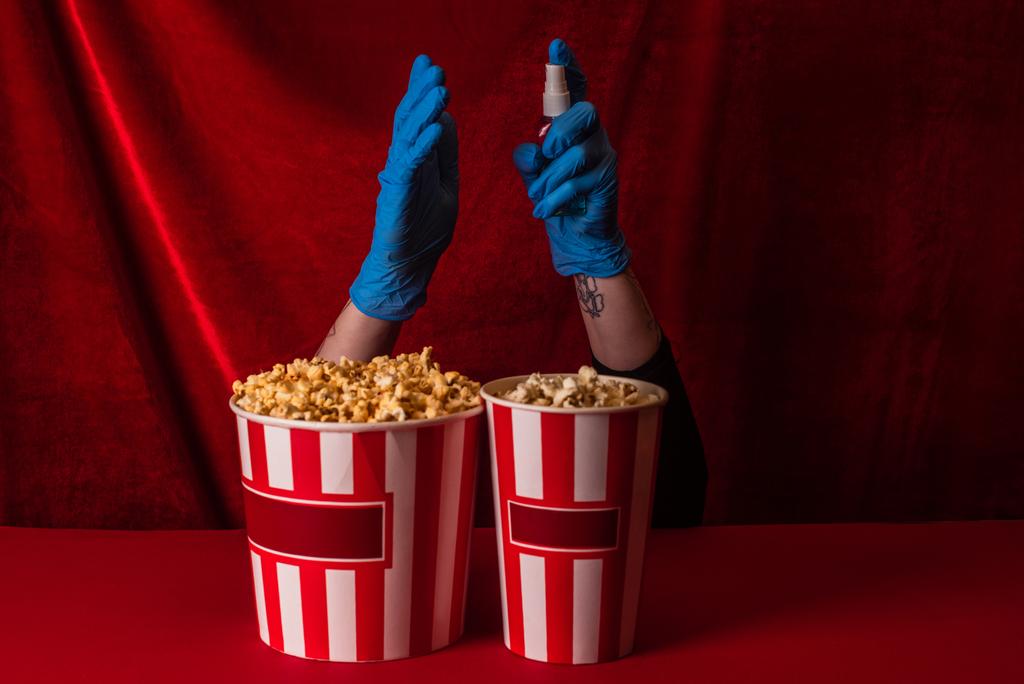 Oříznutý pohled na ženu v latexových rukavicích držící ruční dezinfekci v blízkosti kbelíků s popcornem na červeném povrchu s velurem na pozadí  - Fotografie, Obrázek
