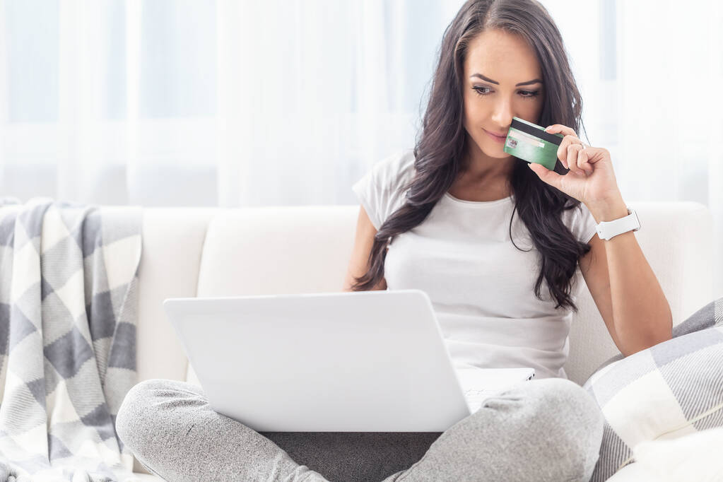 Mujer joven y atractiva considerando comprar en línea mientras está sentada en su sofá blanco con un portátil en su regazo
.  - Foto, Imagen