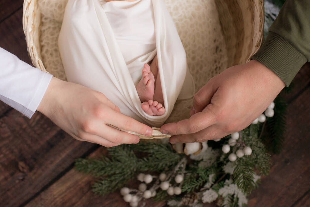 Les mains de la mère et du père tiennent de jolies petites jambes de bébé dans une couverture blanche dans un panier en osier décoré de branches de conifères et de coton sur un sol en bois. Bonne maternité
 - Photo, image