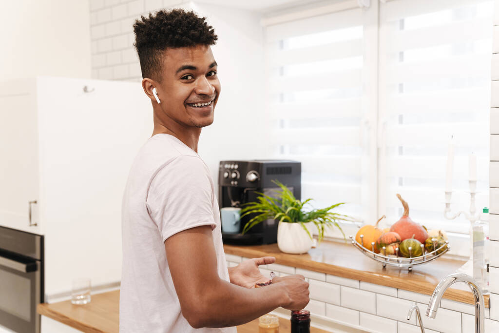 Foto di giovane africano ragazzo positivo al mattino in cucina cucinare la colazione fare panini dolci marmellata ascoltare musica con gli auricolari
. - Foto, immagini