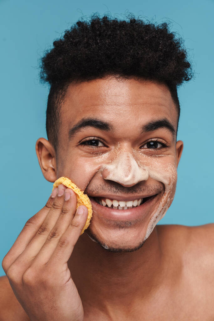 Фотографія безсоромного афро-американського чоловіка, який посміхається, чистячи обличчя губкою і піною, ізольована на синьому фоні. - Фото, зображення