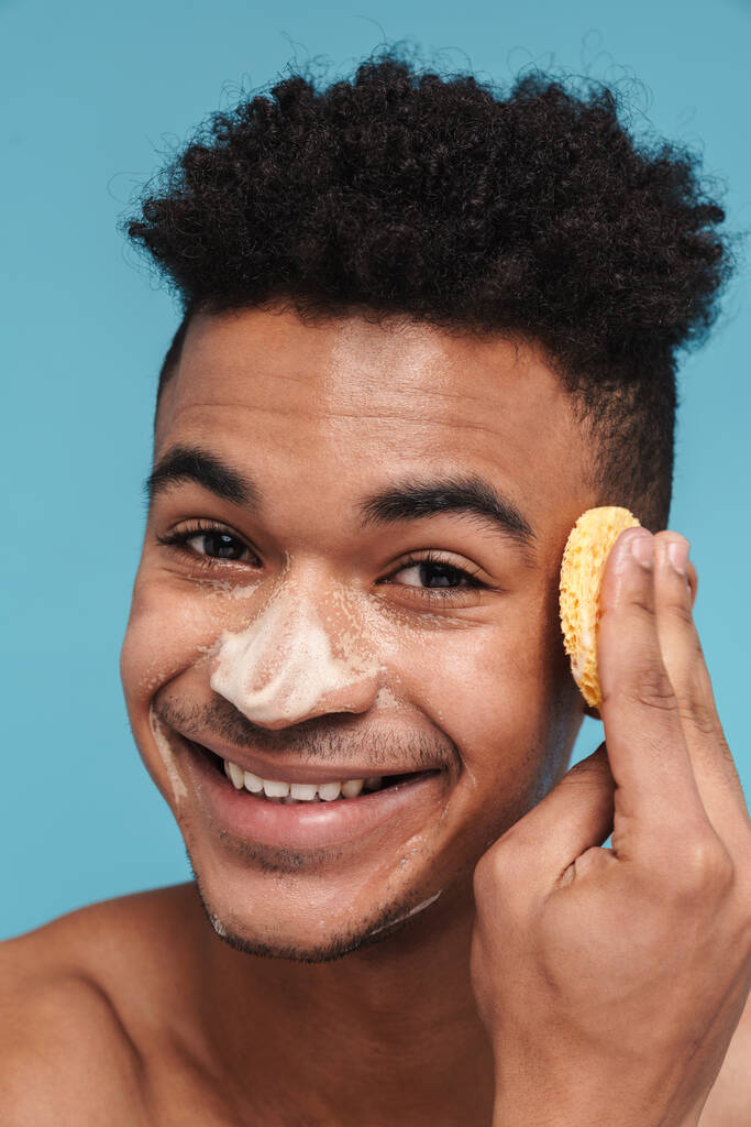 Фотографія безсоромного афро-американського чоловіка, який посміхається, чистячи обличчя губкою і піною, ізольована на синьому фоні. - Фото, зображення