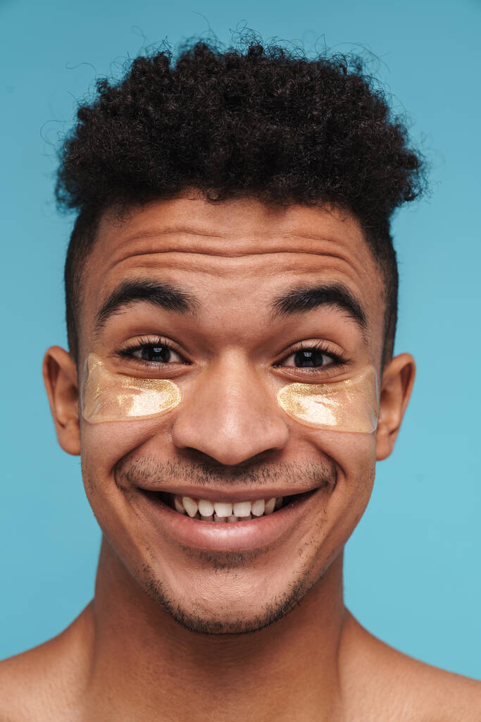 Foto de hombre afroamericano excitado con parches debajo de los ojos sonriendo a la cámara aislada sobre fondo azul
 - Foto, imagen
