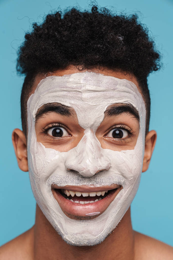 Foto de hombre afroamericano excitado en máscara cosmética facial sonriendo a la cámara aislada sobre fondo azul
 - Foto, imagen