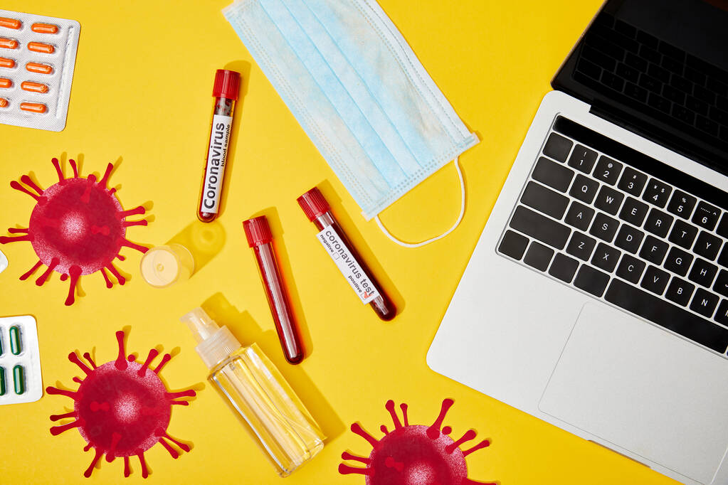 górny widok probówek z napisem koronawirusa w pobliżu maski medycznej, pigułki, butelka z płynem antybakteryjnym i laptop na żółtym  - Zdjęcie, obraz