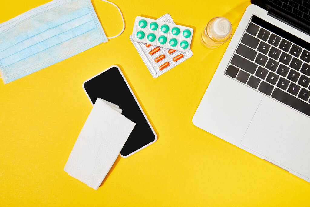 vista superior do laptop e smartphone com tela em branco perto de guardanapo, pílulas, máscara médica e garrafa com higienizador de mão no amarelo
  - Foto, Imagem