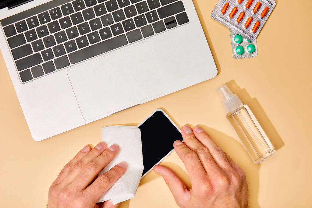 top view of man holding napkin near smartphone with blank screen, laptop, pills and hand sanitizer on beige - Photo, Image
