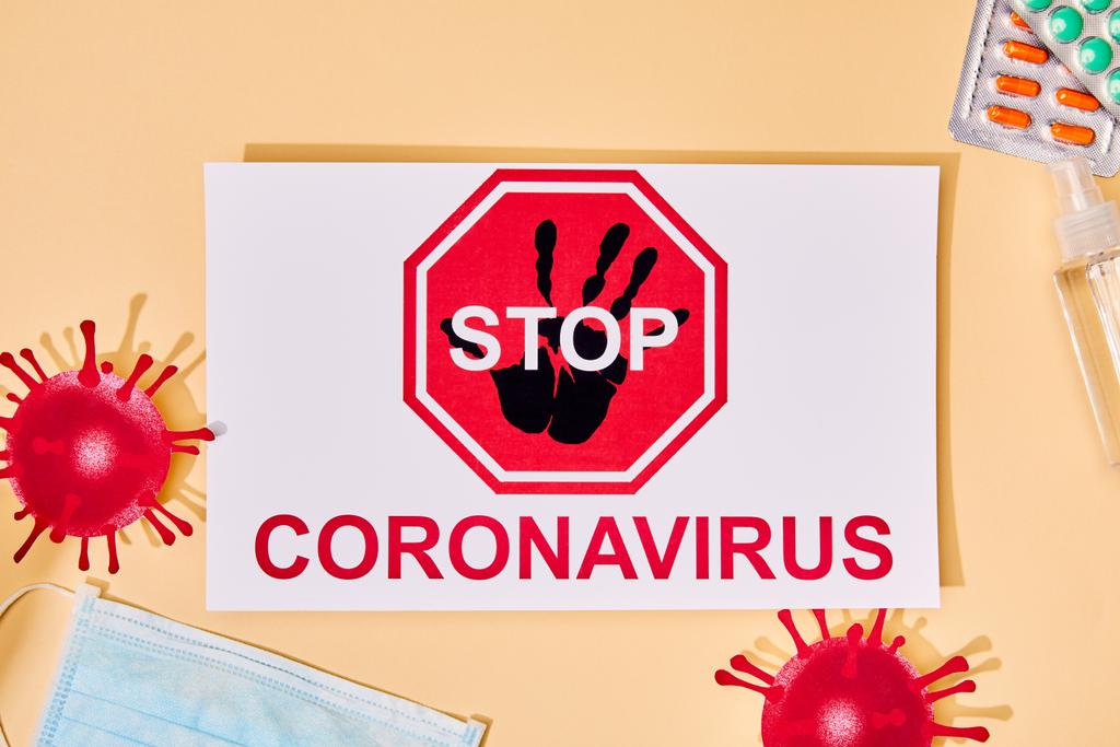 górny widok papieru z literami stop coronavirus w pobliżu wyciągniętego wirusa, maska medyczna, dezynfektor rąk i pigułki na beżu  - Zdjęcie, obraz
