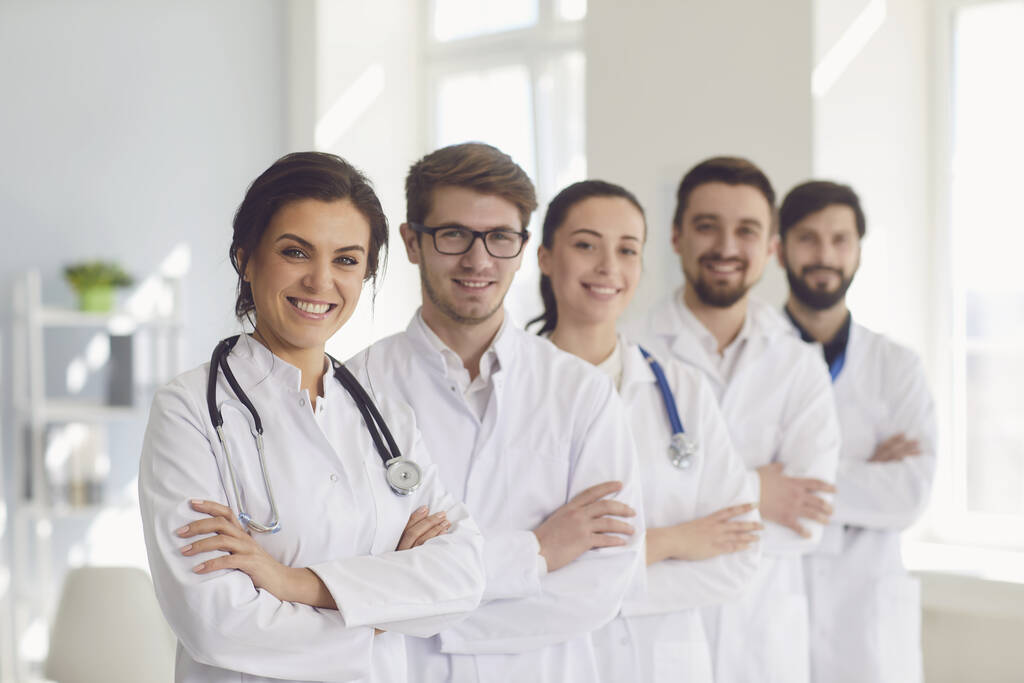 A group of confident practicing doctors in white coats are smiling against the backdrop of the clinic. - Photo, Image