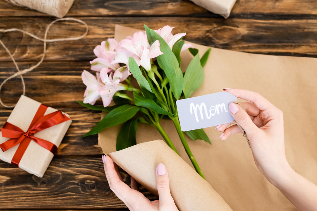 bovenaanzicht van vrouw met label met moeder belettering in de buurt van roze bloemen en geschenkdoos op houten oppervlak, Moederdag concept  - Foto, afbeelding