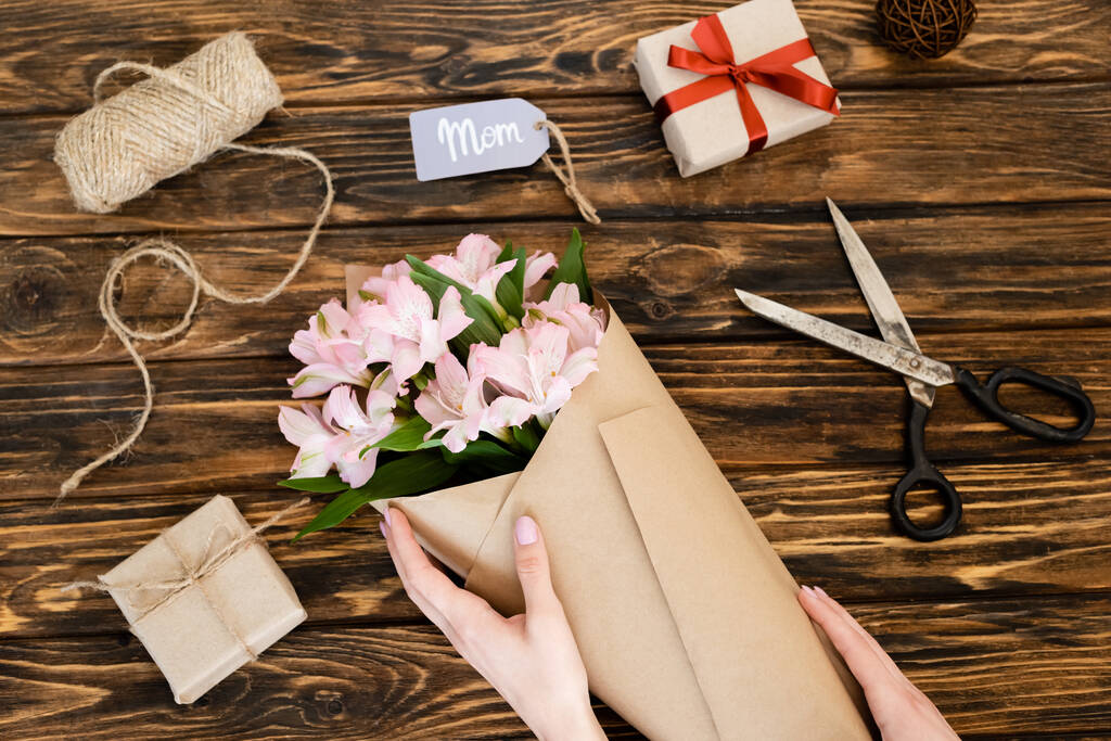 vue recadrée de femme touchant enveloppé dans des fleurs roses en papier près de boîtes-cadeaux et tag avec maman lettrage sur la surface en bois, concept de fête des mères
  - Photo, image