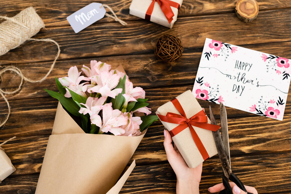 cropped view of woman cutting ribbon on present near pink flowers, greeting card with happy mothers day and tag with mom lettering on wooden surface - Photo, Image