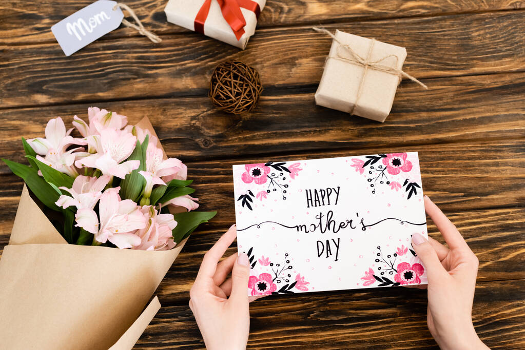 vista recortada de la mujer que sostiene la tarjeta de felicitación con letras feliz día de las madres cerca de flores y regalos en la superficie de madera
  - Foto, Imagen
