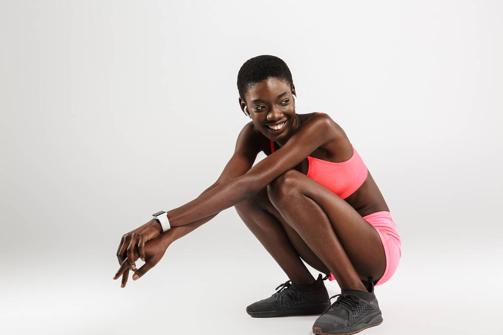 Obrázek usmívající se africké americké sportovkyně pomocí bezdrátových sluchátek při dřepění izolované nad bílým pozadím - Fotografie, Obrázek