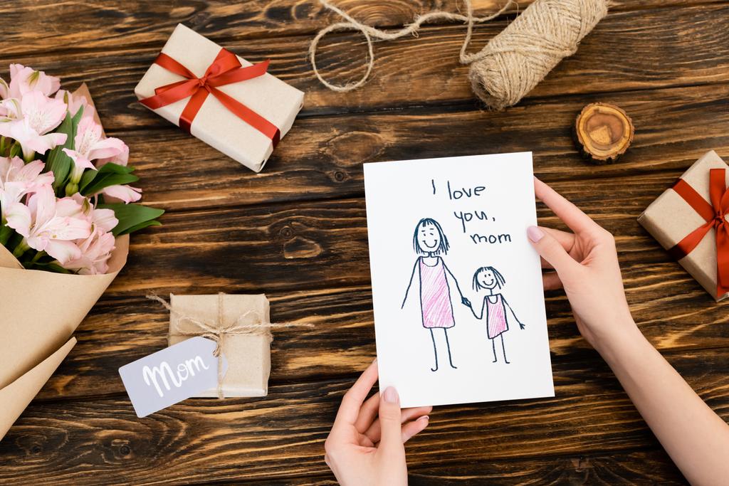 abgeschnittene Ansicht einer Frau mit Grußkarte mit i love you mom Schriftzug in der Nähe von Geschenken und Blumen auf hölzerner Oberfläche - Foto, Bild