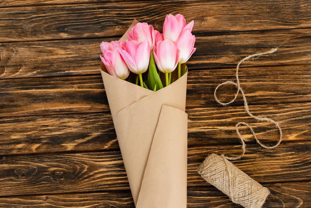 widok z góry różowe tulipany zawinięte w papier w pobliżu jutowej sznurka liny na drewnianej powierzchni, koncepcja dzień matki  - Zdjęcie, obraz