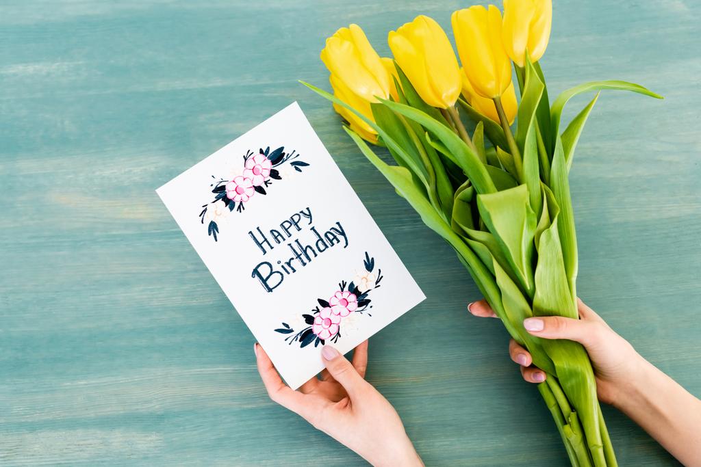 vista recortada de la mujer que sostiene la tarjeta de felicitación con letras de feliz cumpleaños y tulipanes amarillos en la superficie de madera azul
  - Foto, Imagen