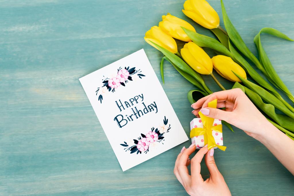 vista ritagliata della donna che tiene la scatola regalo vicino al biglietto di auguri con scritte di buon compleanno e tulipani gialli sulla superficie di legno blu
  - Foto, immagini