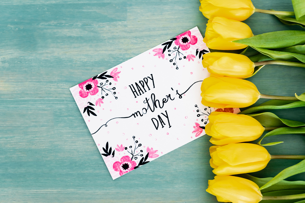 top view üdvözlőlap boldog anyák napi felirat közelében sárga tulipán kék texturált felületen - Fotó, kép