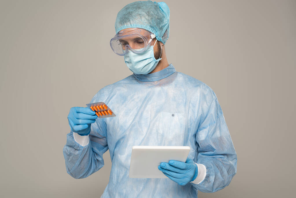 Lékař v lékařské masce drží blistr s pilulkami a digitální tablety izolované na šedé - Fotografie, Obrázek