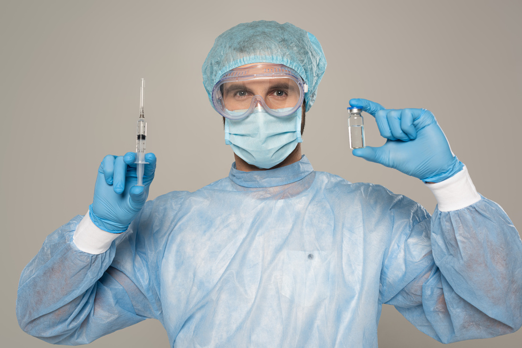 Γιατρός σε προστατευτικό googles και ιατρική μάσκα κρατώντας σύριγγα και βάζο του εμβολίου που απομονώνονται σε γκρι - Φωτογραφία, εικόνα