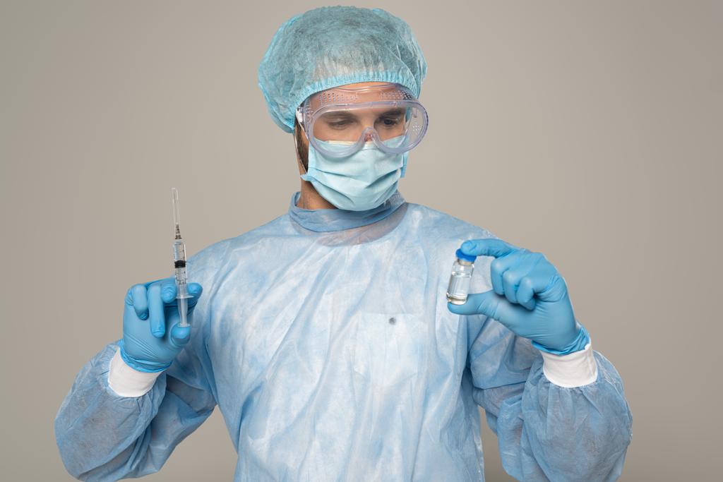 Lekarz w masce medycznej i ochronnych google trzymając słoik szczepionki i strzykawki izolowane na szaro - Zdjęcie, obraz