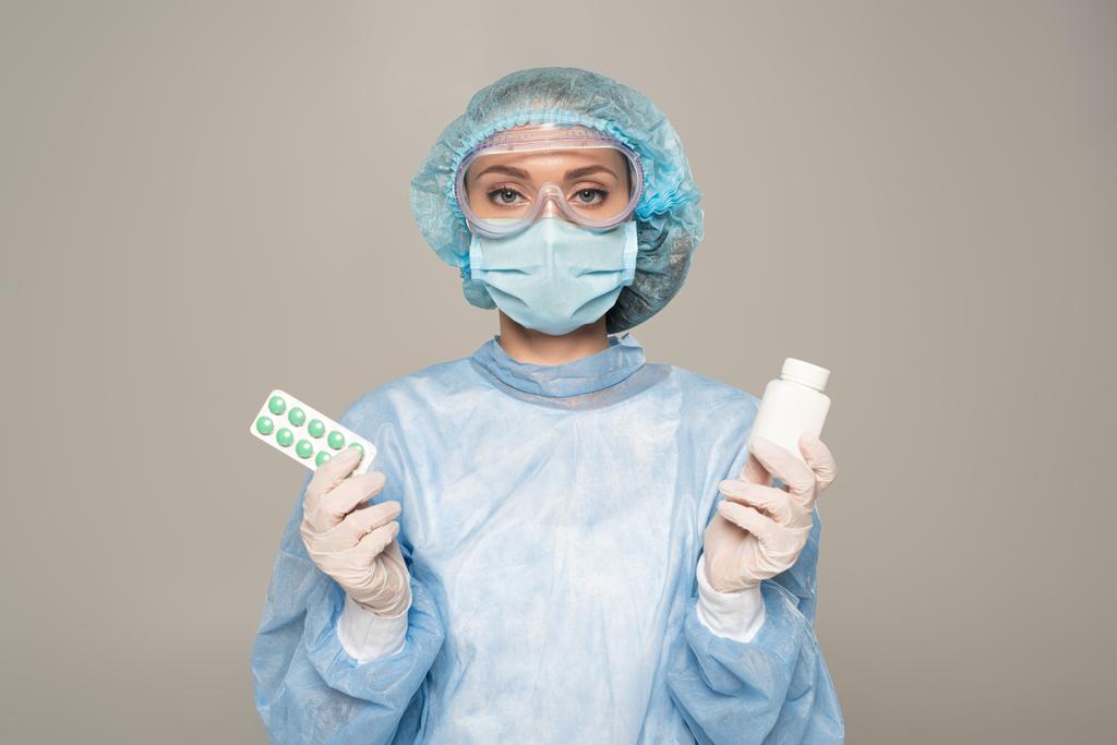 Doctor in medical mask and protective googles holding blister and jar of pills isolated on grey - Photo, Image