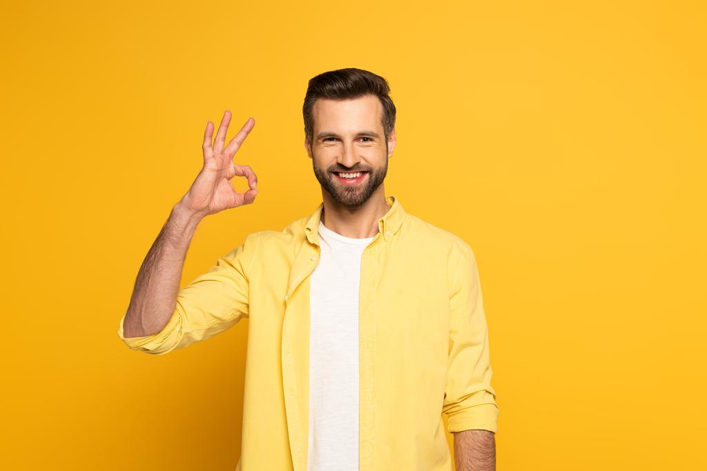 Όμορφος άνθρωπος χαμογελά στην κάμερα, ενώ δείχνει ok σημάδι στο κίτρινο φόντο - Φωτογραφία, εικόνα