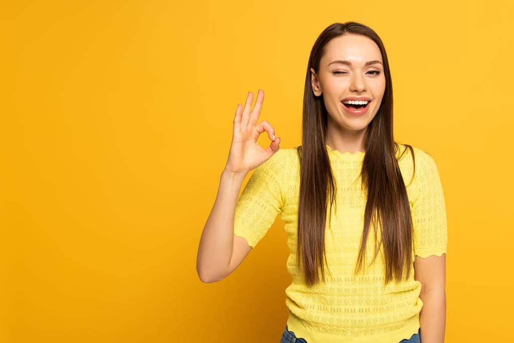 Attraente sorridente ragazza ammiccante mentre mostra ok gesto su sfondo giallo
 - Foto, immagini