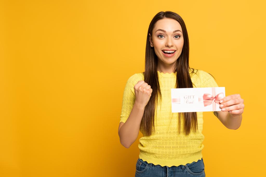Mujer feliz sosteniendo la tarjeta de regalo y mostrando sí gesto sobre fondo amarillo
 - Foto, Imagen
