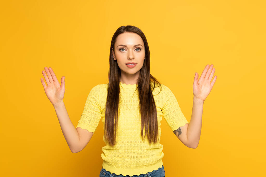 Mujer joven mirando a la cámara mientras usa lenguaje de señas sobre fondo amarillo
 - Foto, Imagen