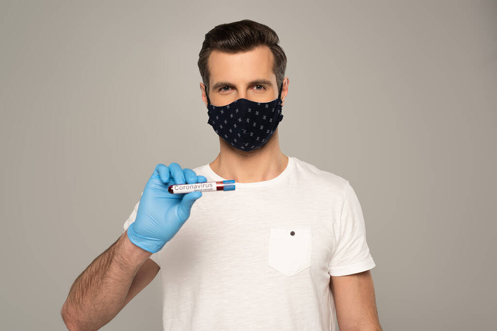 Man in safety mask and latex glove holding test tube with blood sample and coronavirus lettering isolated on grey  - Photo, Image