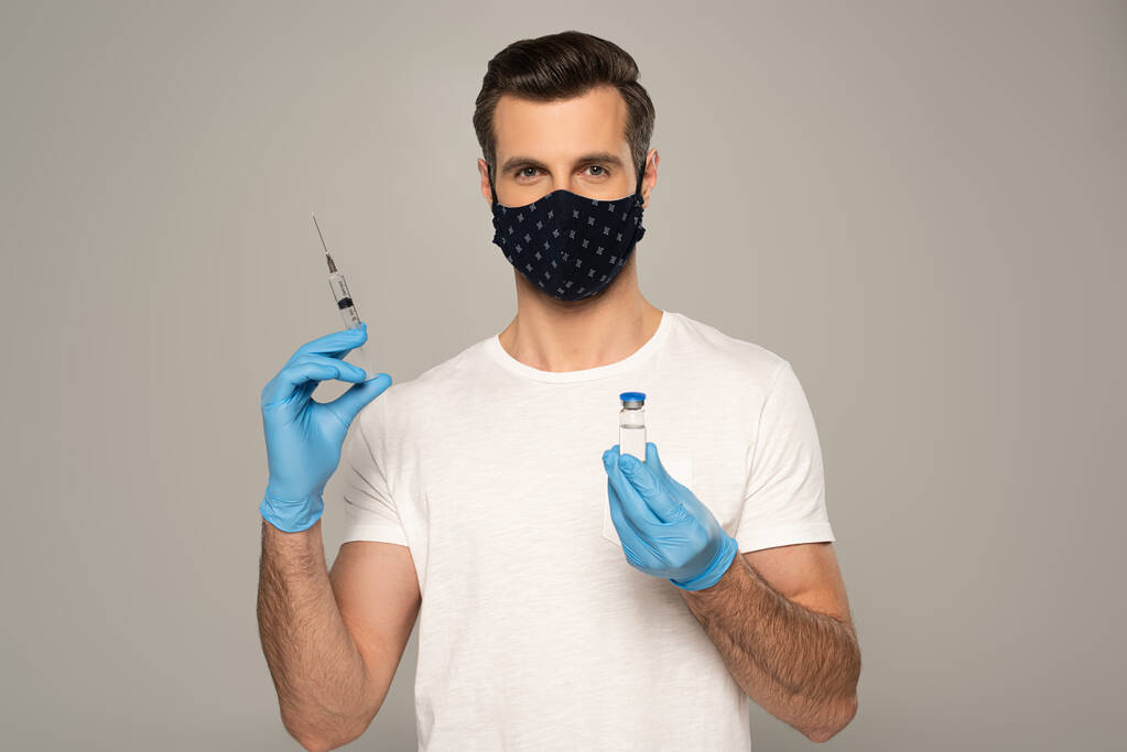 Homem com máscara de segurança e luvas de látex segurando frasco de vacina e seringa isolado em cinza
  - Foto, Imagem