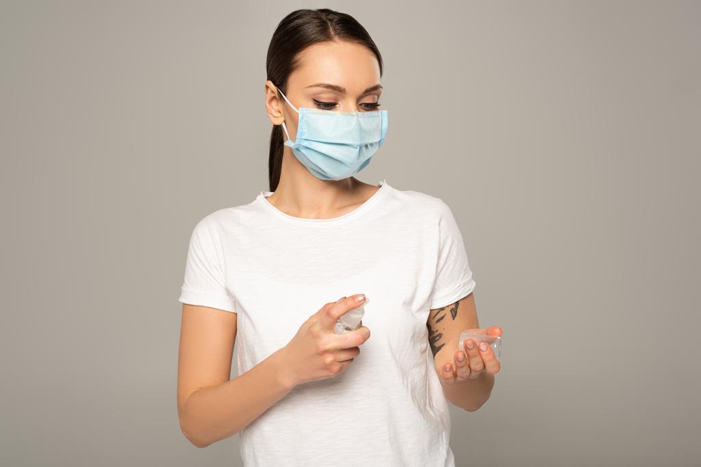 Молода жінка в медичній масці використовує дезінфікуючий засіб для рук ізольовано на сірому
  - Фото, зображення