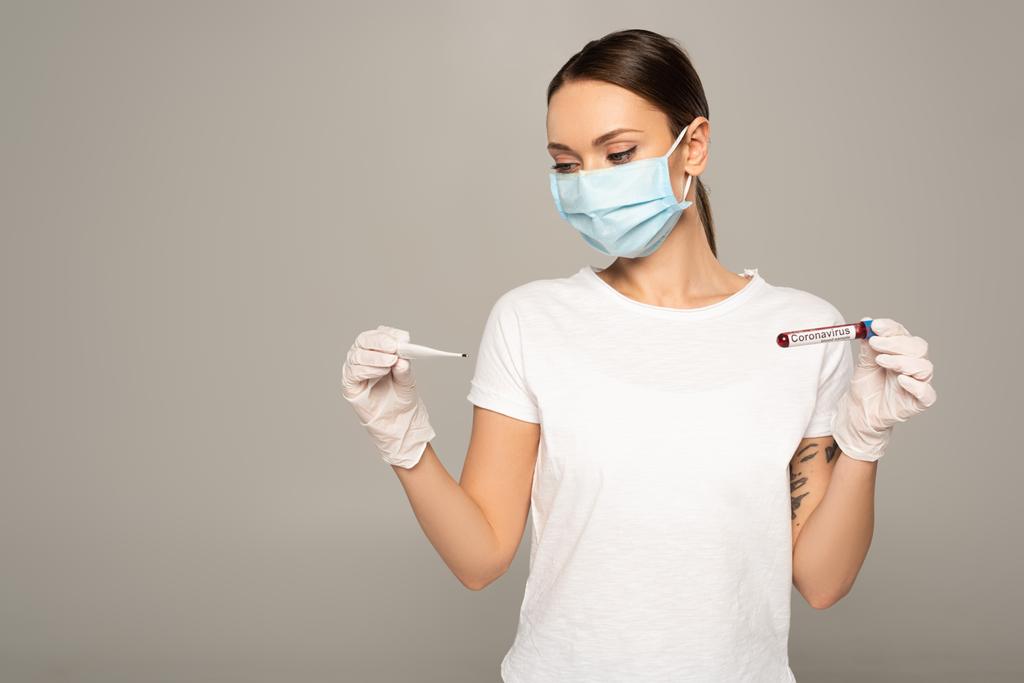 Junge Frau in medizinischer Maske hält Thermometer und Reagenzglas mit Blutprobe und Coronavirus-Schriftzug isoliert auf grau  - Foto, Bild