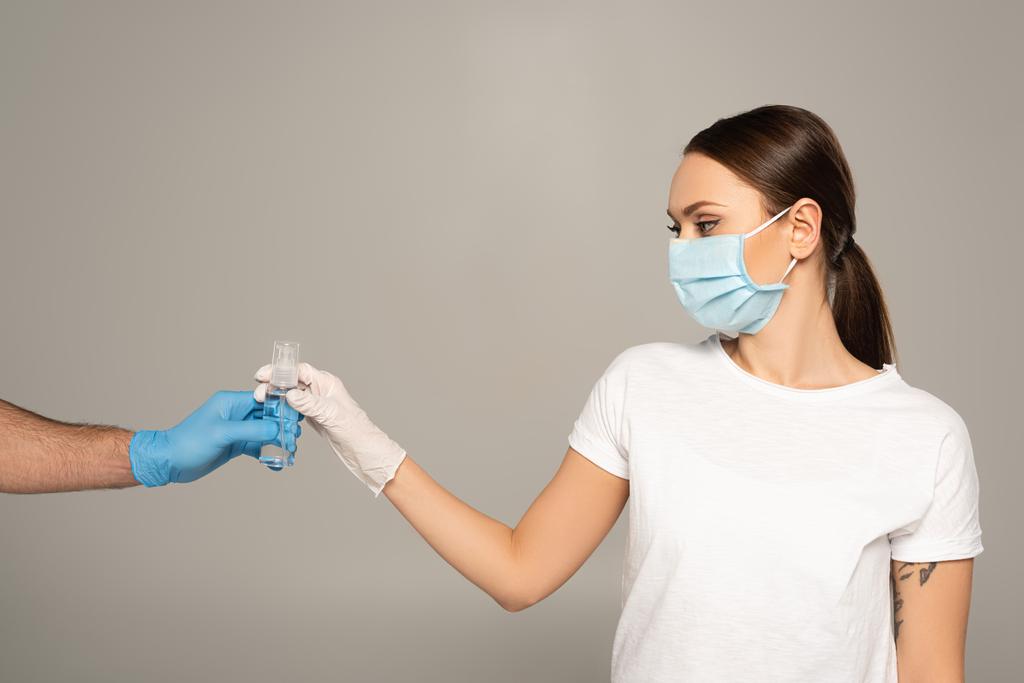 Vrouw met medisch masker neemt fles handreiniger van man in latex handschoen geïsoleerd op grijs  - Foto, afbeelding