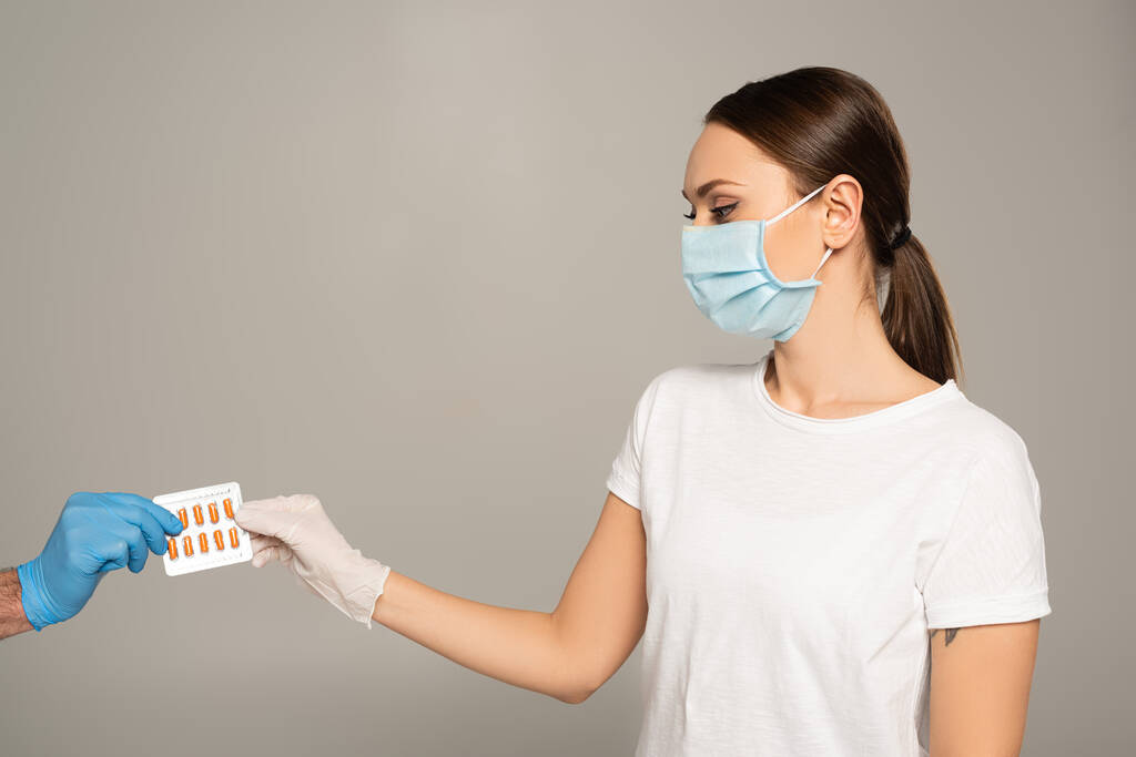 Donna in maschera medica con blister con pillole vicino all'uomo in guanto di lattice isolato su grigio
  - Foto, immagini