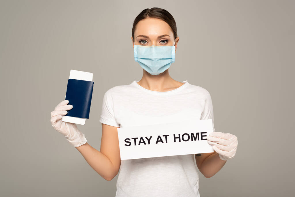 Женщина в медицинской маске, держащая паспорт с авиабилетом и карточкой с пребыванием дома, надпись изолирована на сером
  - Фото, изображение